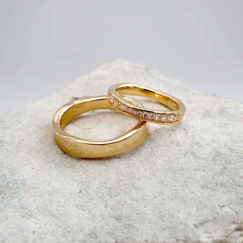 To vielsesringe i guld - en til manden og en til kvinden. Kvindens ring har diamanter langt toppen af ringen. Ringene ligger på en lys sten. 